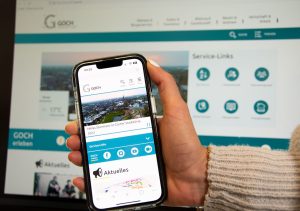 Hand mit Smartphone vor einem Bildschirm. Zu sehen ist die neue Website der Stadt Goch, bei der auch der Mängelmelder Goch prominent platziert und verlinkt ist.