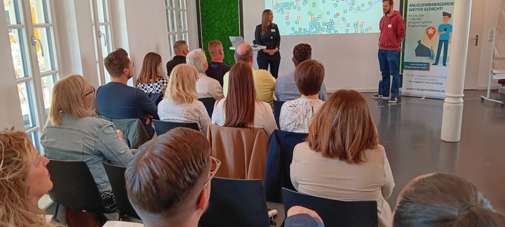Vortrag beim Info- und Anwendertag Mängelmelder 2025. Die Referenten stehen vor der Leinwand, die Teilnehmer sitzen in Stuhlreihen und hören zu.