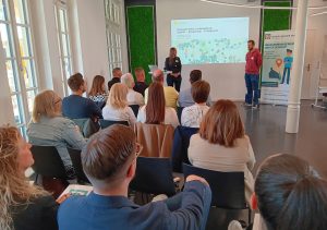 Vortrag beim Info- und Anwendertag Mängelmelder 2025. Die Referenten stehen vor der Leinwand, die Teilnehmer sitzen in Stuhlreihen und hören zu.