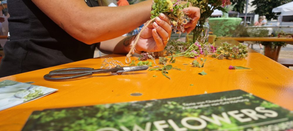 Tisch mit Blumen; Person beim Binden eines Blumenkranzes im Rahmen vom Aktionstag "Mit(te) gemacht" in der Stadt Griesheim.