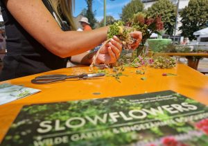 Tisch mit Blumen; Person beim Binden eines Blumenkranzes im Rahmen vom Aktionstag "Mit(te) gemacht" in der Stadt Griesheim.