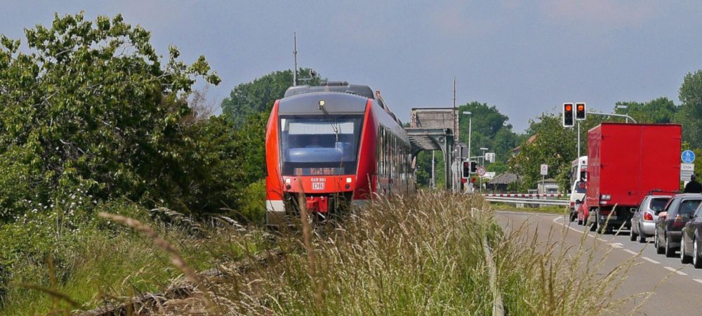 Regionalzug in Richtung Kiel fährt neben einer Straße mit einer Autoschlange. Symbolbild für den landesweiten Nahverkehrsplan Schleswig-Holstein, zu dem im September 2025 Online-Foren mit den relevanten Stakeholdern stattgefunden haben.