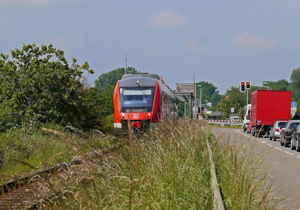 Regionalzug in Richtung Kiel fährt neben einer Straße mit einer Autoschlange. Symbolbild für den landesweiten Nahverkehrsplan Schleswig-Holstein, zu dem im September 2025 Online-Foren mit den relevanten Stakeholdern stattgefunden haben.